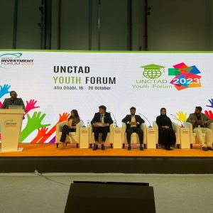 Fotografía del escenario del evento. De izquierda a derecha, se encuentra el moderador del espacio ubicado en un palco, luego, sentados en sillones, se encuentra Gerónimo junto a cuatros jóvenes, hombres y mujeres, que conversan y exponen sus ideas al público. Detrás, una pantalla gigante con los logos del World Ivestment Forum 2023 y del Unctad Youth Forum. ‌