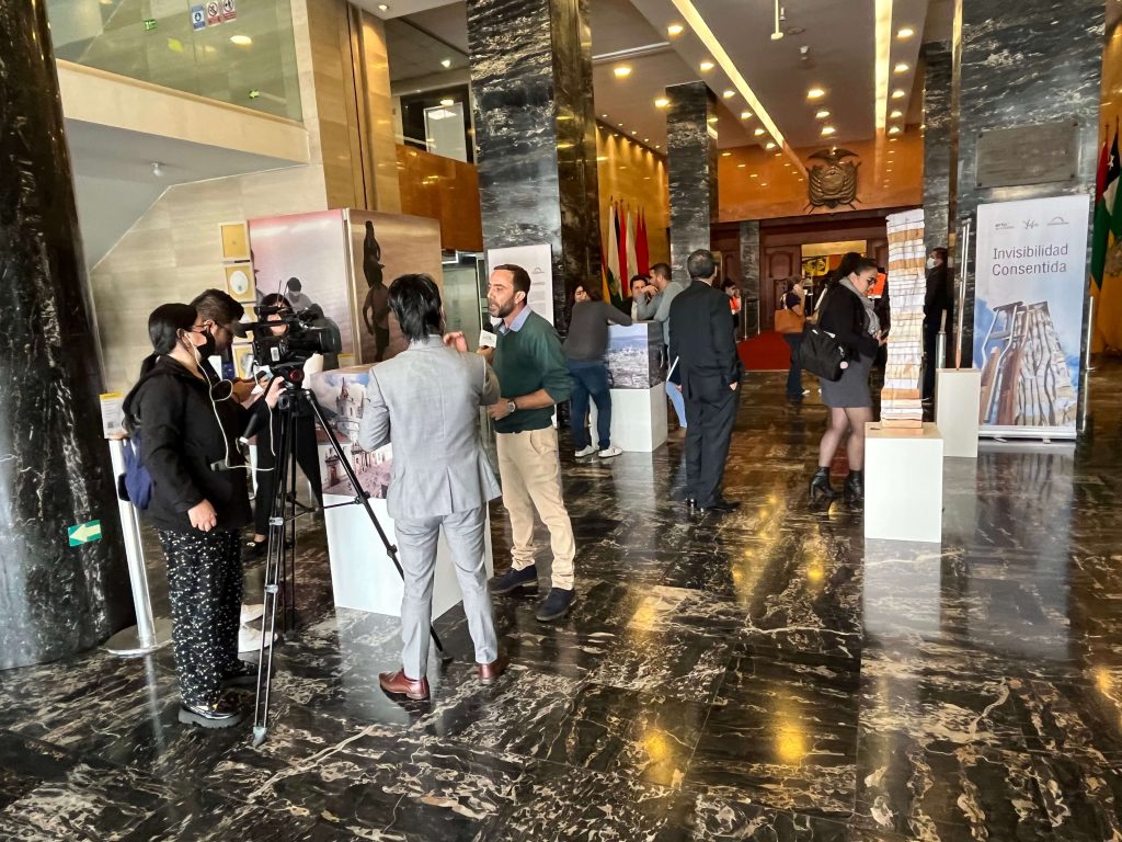 Photo of Andrés Julio addressing journalists at the exhibition. Behind him, there are some pieces of art and various people observing them. Besides, there is a banner with the name of the exhibition: "Invisibilidad Consentida".