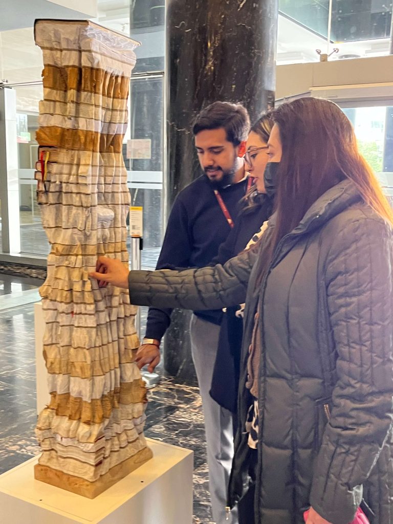 Foto de los visitantes de la muestra tocan con sus manos una obra de arte llamada "Soporte Legal 3". Esta obra consiste en varias constituciones apiladas, formando una torre de aproximadamente 1,5 metros.