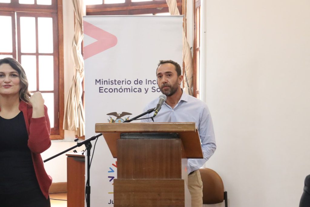 Andrés Julio, artist responsible for the exhibition, makes his presentation addressing the audience, standing on a plataform. Behind him, there is a banner that says “Ministry of Economic and Social Inclusion”. Beside Andrés, there is a Sign Language interpretation.