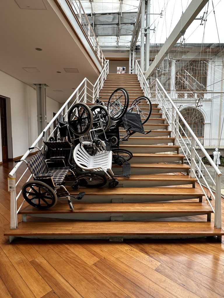 Photo of a staircase featuring the artwork "In-movilidad" which consists of several chairs and wheelchairs stacked together, representing the redefinition of mobility spaces and tools.