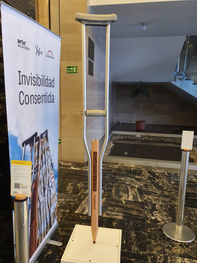 Foto de la obra “Soporte Legal #1”, exhibida en la muestra Invisibilidad Consentida, en Quito, Ecuador. Detrás de la obra hay un banner en el que se lee el nombre de la muestra, acompañado por el logo del Programa Arte Accesible, la firma de Andrés Julio - artista a cargo de la muestra - y el logo de Fundación Comparlante.