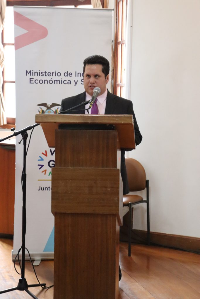 Foto de Sebastián Flores de pie, realizando una presentación dirigiéndose al público presente, parado sobre una tarima. Detrás de Sebastian, hay un banner del Ministerio de Inclusión Económica y Social.